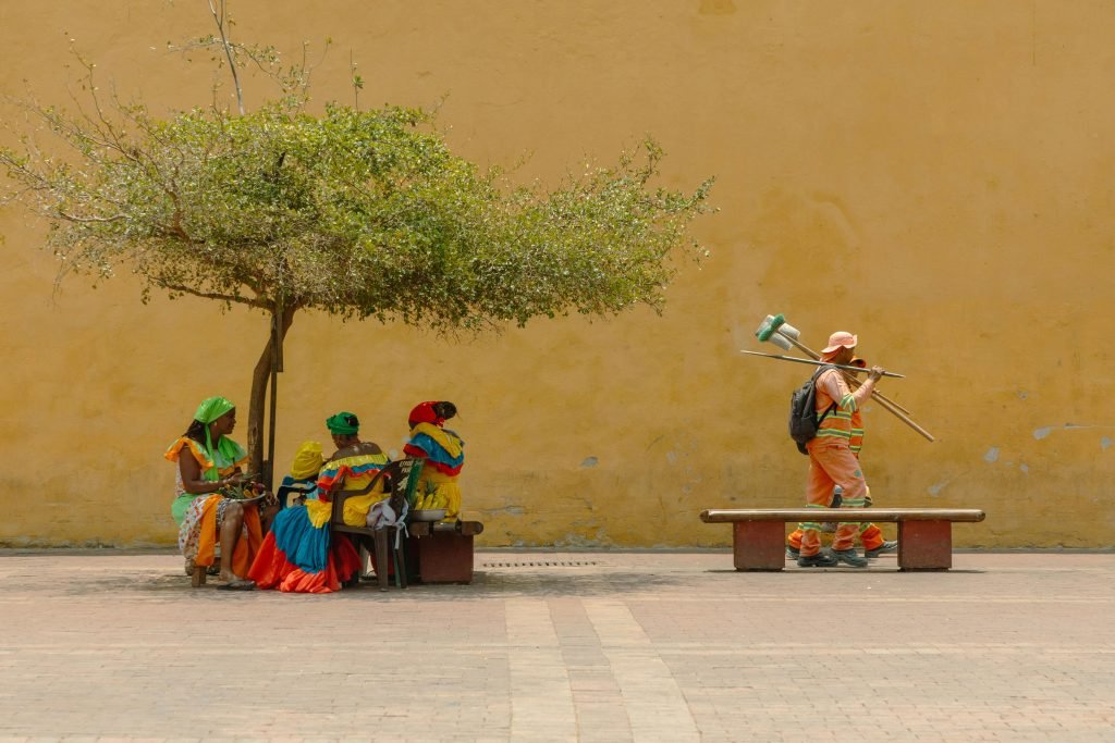 Women in vibrant traditional attire rest under a tree while a worker passes by, viajes a Cartagena
