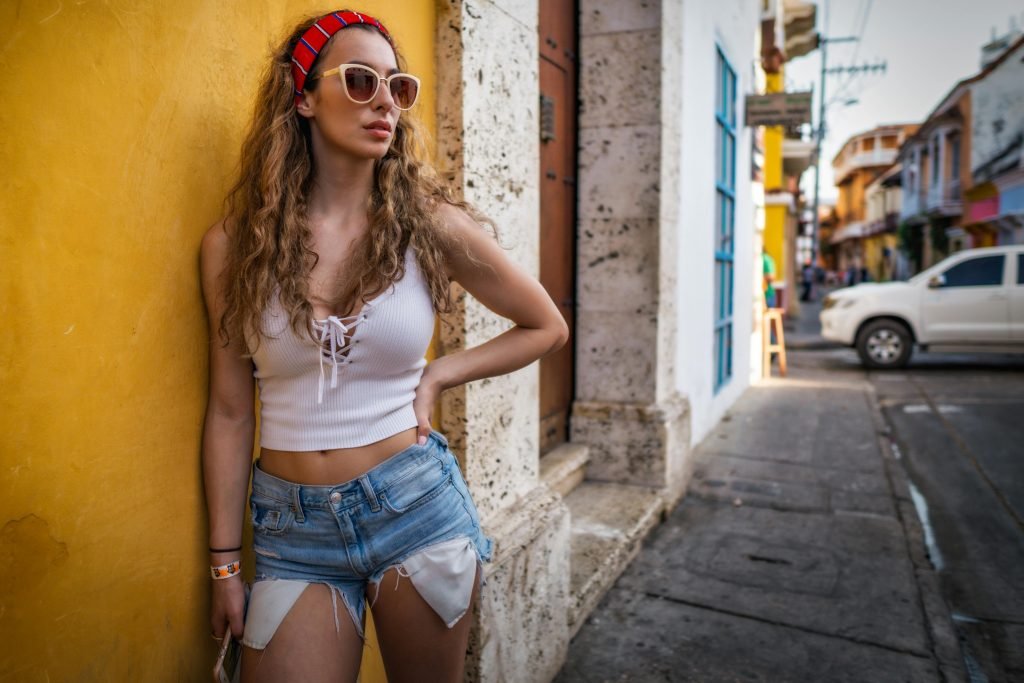 A fashionable woman in a white top and denim shorts leans against a colorful wall in Cartagena, Colombia, viajes a Cartagena