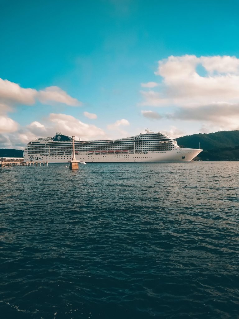 A grand cruise ship sails under a bright blue sky with fluffy clouds over a calm ocean, cruceros