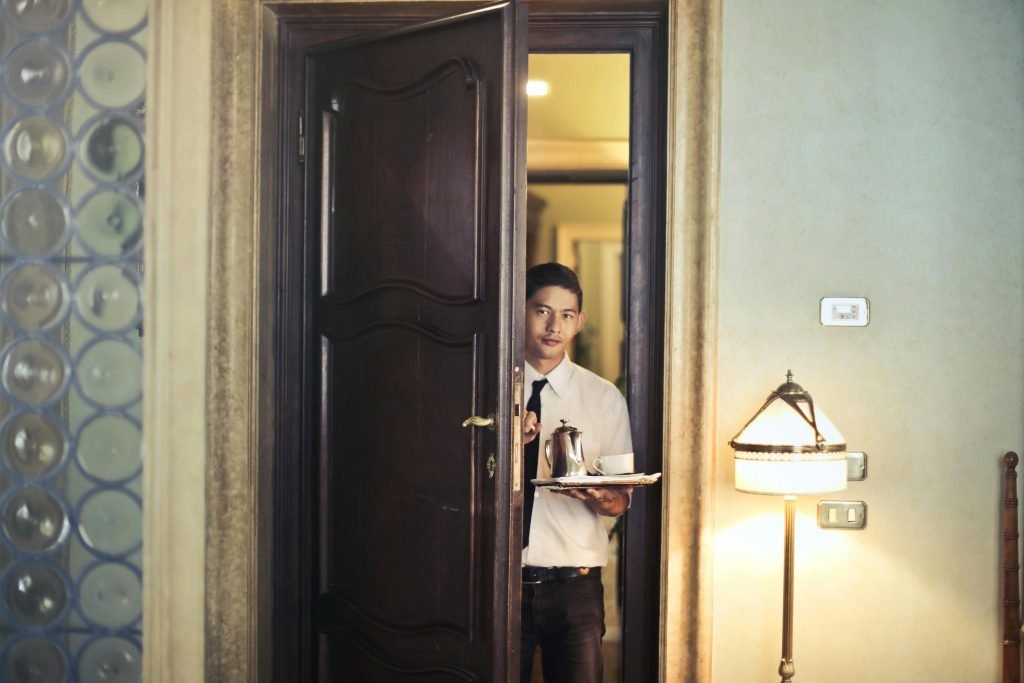 Young ethnic male room service waiter carrying tray with coffee pot while entering hotel room with stylish vintage interior, hotelería