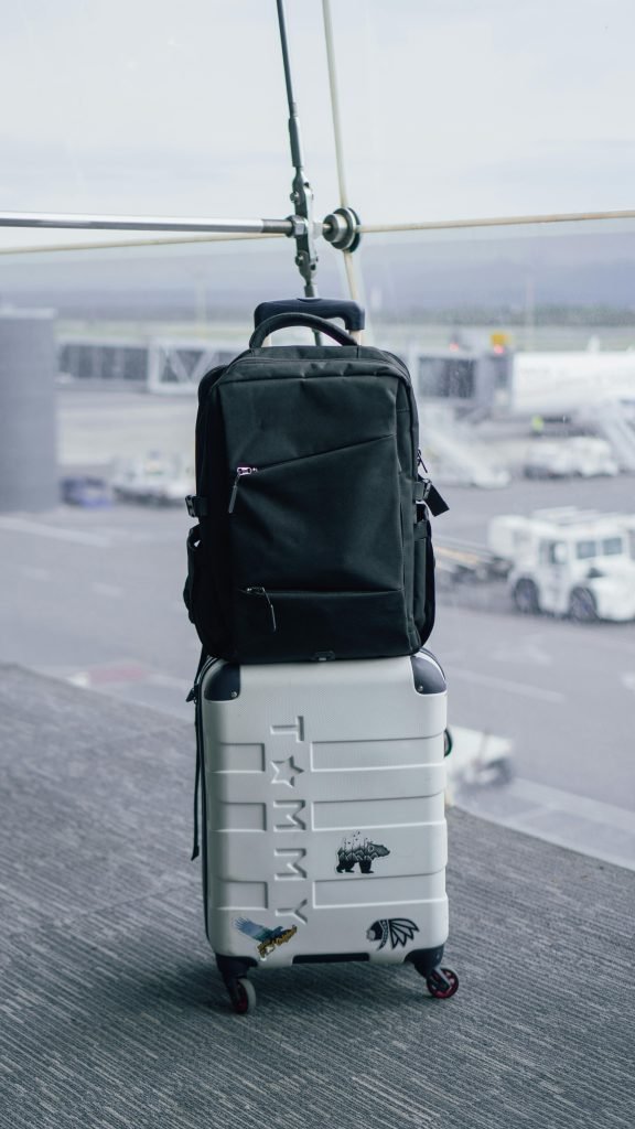 A black backpack sits atop a white suitcase in an airport terminal with a tarmac view., equipaje