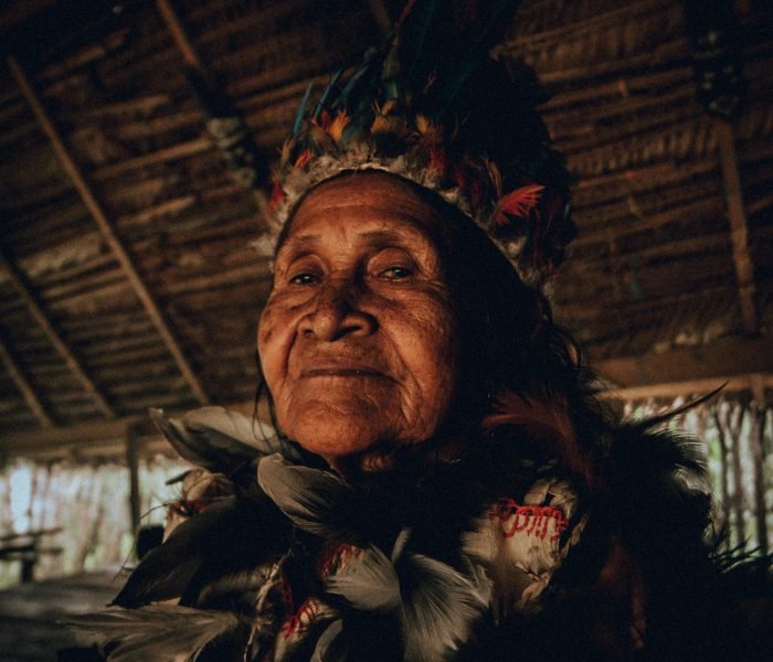 Elderly indigenous woman wearing traditional feather headdress in an Amazonian hut.