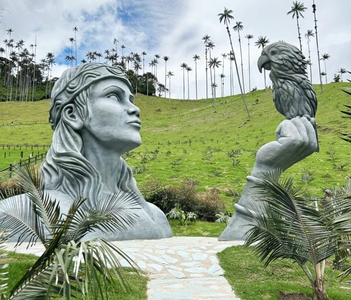 Large stone sculptures in lush landscape of Salento, featuring a human and bird amidst tall palm trees.